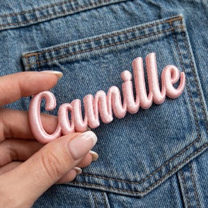 Könnte beinhalten: Ein hellrosa gestickter Aufnäher mit dem Namen Camille in Schreibschrift. Die Buchstaben haben eine erhabene, gepolsterte Optik mit einer glatten, satinartigen Oberfläche und sind für die Anbringung auf Stoffen wie Jeans gedacht.