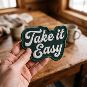 May include: A dark green embroidered patch with a scalloped edge featuring the white stitched text Take it Easy in a cursive font. The patch has a thick green border and a textured fabric surface, designed for application onto various textile items.
