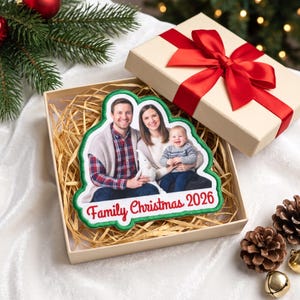 Puede incluir: Un adorno de forma personalizada con una fotografía impresa de una familia de tres personas, con el texto rojo Family Christmas 2026 en la parte inferior. El adorno tiene un borde verde y está en una caja de regalo con lazo rojo.