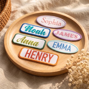 Op de afbeelding: Een verzameling van zes geborduurde naamlabels in rechthoekige en ovale vormen. De labels zijn gemaakt van witte stof met gekleurde randen en bevatten de namen Sophia, Noah, James, Anna, Emma en Henry in diverse lettertypes en kleuren.