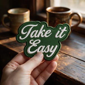 Puede incluir: Un parche bordado de color verde oscuro con un borde festoneado que presenta el texto blanco Take it Easy en una fuente cursiva. El parche tiene un borde cosido grueso y una textura de tela, mostrando las palabras claramente en la superficie.