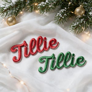 Peut inclure: Deux écussons brodés affichant le prénom Tillie dans une police cursive. Un écusson est cousu avec du fil rouge vif, tandis que l'autre est cousu avec du fil vert foncé. Les deux écussons présentent une bordure blanche et un relief texturé.