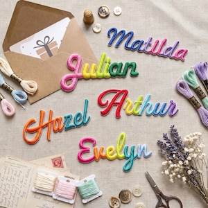 Könnte beinhalten: Fünf bestickte Namensaufnäher in Schreibschrift mit den Namen Matilda, Julian, Hazel, Arthur und Evelyn in leuchtenden Farbverläufen. Die Aufnäher sind mit Nähzubehör wie Stickgarn, Knöpfen und einem braunen Umschlag arrangiert.