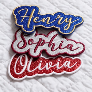 May include: Three custom name patches with glittery backgrounds and embroidered script lettering. The top patch is blue with gold text reading Henry, the middle is silver with maroon text reading Sophia, and the bottom is red with white text reading Olivia.
