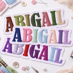 Puede incluir: Tres parches bordados con el nombre Abigail en varios colores. El parche superior tiene letras en marrón, oliva, verde oscuro, rosa viejo y beige. El parche central presenta letras en azul marino, rosa, menta, azul claro, melocotón y lavanda.