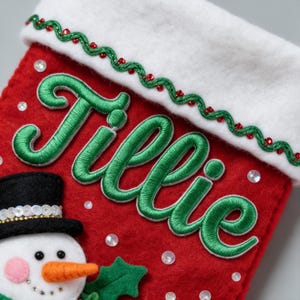 以下が含まれることがあります： 緑色の筆記体で「Tillie」と刺繍された赤いフェルト製のクリスマスストッキング。白いカフには緑色のジグザグ模様のトリムと赤いスパンコールが施され、黒いシルクハットをかぶった雪だるまのアップリケと、散りばめられた白いスパンコールが特徴です。