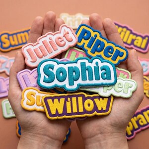 May include: A collection of embroidered name patches featuring various colors and fonts. Visible names include Sophia in teal, Juliet in pink, Piper in blue, and Willow in yellow. The patches have scalloped edges and are layered together.