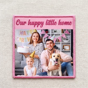 Puede incluir: Un parche de tela cuadrado con borde rosa que presenta una fotografía impresa de una familia celebrando un cumpleaños. El texto "Our happy little home" está bordado en rosa en la parte superior. La imagen muestra a un niño, una mujer y un hombre con un perro.