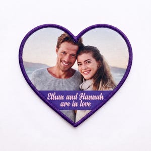 Puede incluir: Un parche de tela en forma de corazón con un borde bordado en color morado. El centro presenta una fotografía impresa de una pareja sonriente. Debajo de la foto, una banda morada contiene el texto en blanco: Ethan and Hannah are in love.