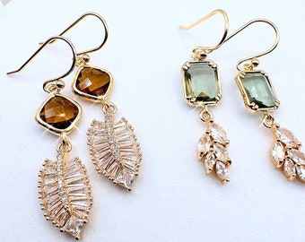 May include: A pair of gold earrings with a leaf design and a square-shaped gemstone. One earring has a brown gemstone and the other has a green gemstone.