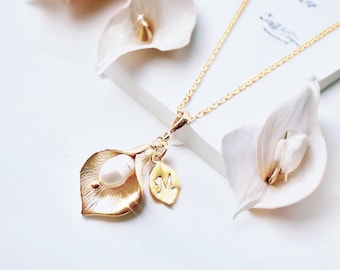Puede incluir: Un collar dorado con un colgante de hoja y perla, con un pequeño dije de hoja. El collar se exhibe con flores de cala blancas. El fondo es blanco.