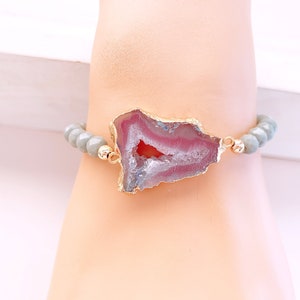 May include: A beaded bracelet featuring a gold-plated, irregularly shaped agate stone with pink and white hues. The bracelet has light gray beads and gold accents, creating a unique and elegant accessory.