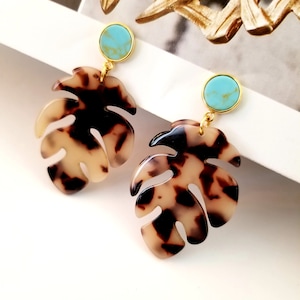 Puede incluir: Un par de pendientes de tono dorado con una piedra turquesa y un diseño de hoja con estampado marrón y blanco.