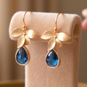 May include: Gold-toned drop earrings with a floral design. Each earring features a cluster of three leaves at the top and a teardrop-shaped, deep blue gemstone. The earrings are displayed on a beige velvet jewelry stand.