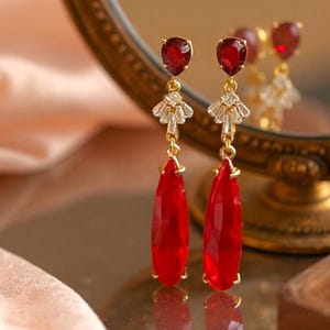 May include: A pair of elegant drop earrings featuring teardrop-shaped, vibrant red gemstones. The earrings have a gold-colored setting with smaller red stones at the top and a decorative element of clear crystals. The earrings are reflected in a gold-framed mirror.
