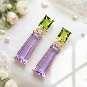 Può includere: Un paio di orecchini pendenti con una pietra preziosa rettangolare sfaccettata di colore viola sospesa sotto una pietra quadrata sfaccettata di colore verde. Le pietre sono incastonate in metallo dorato con un piccolo gruppo di cristalli chiari.