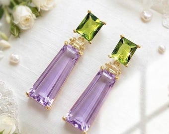 May include: A pair of gold-tone drop earrings featuring a rectangular, faceted purple stone suspended beneath a smaller, rectangular, faceted lime green stone. The earrings are set in a gold-colored metal frame with a polished finish.