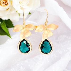 May include: Gold-toned earrings featuring a floral design with three petals and a teardrop-shaped, emerald-green gemstone. The earrings are set against a white background with white roses.