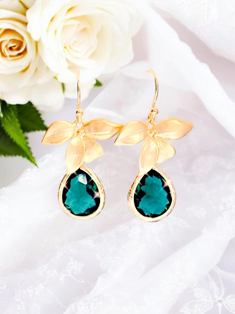 May include: Gold-toned earrings featuring a floral design with three petals and a teardrop-shaped, emerald-green gemstone. The earrings are set against a white background with white roses.