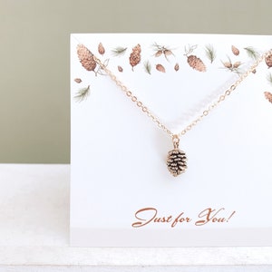 May include: Gold necklace with a small pine cone pendant. The necklace is displayed on a white card with a pine cone design and the text "Just for You!"