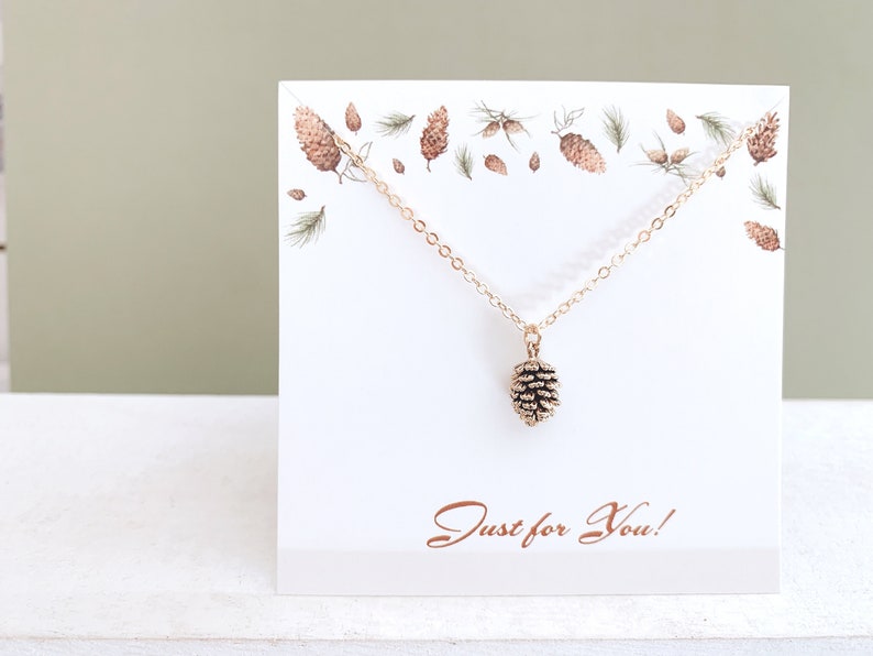 May include: Gold necklace with a small pine cone pendant. The necklace is displayed on a white card with a pine cone design and the text "Just for You!"