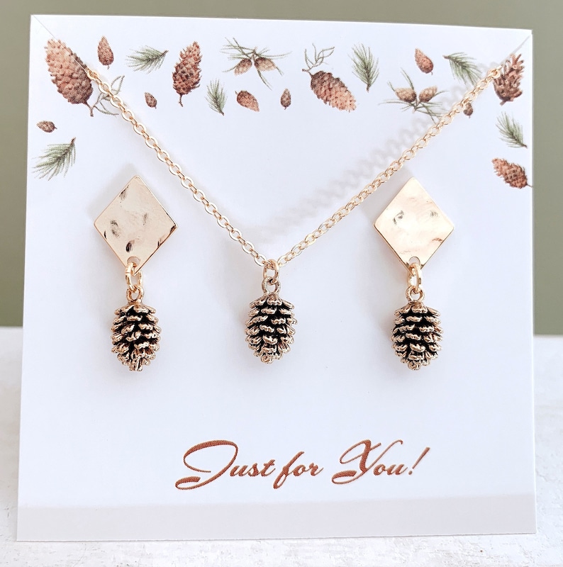 May include: A gold necklace and earrings set featuring a pine cone pendant and matching earrings. The necklace and earrings are displayed on a white card with the text "Just for You!" printed on it.
