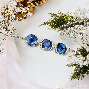 May include: A delicate gold chain bracelet with three square-shaped blue crystal stones. The bracelet is on a white background with white and green flowers.