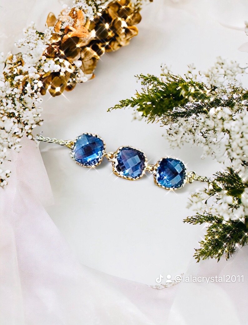May include: A delicate gold chain bracelet with three square-shaped blue crystal stones. The bracelet is on a white background with white and green flowers.