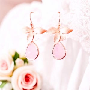 May include: Rose gold floral earrings with teardrop-shaped pink gemstones. The earrings feature a delicate flower design with multiple petals and a hook closure. The gemstones are a soft pink color.