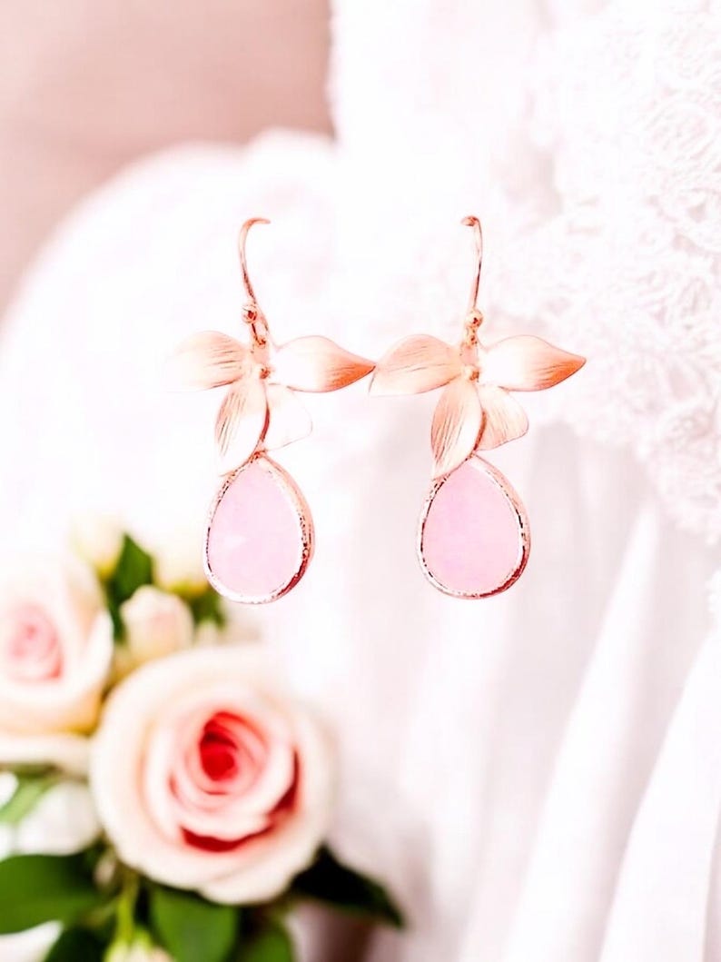 May include: Rose gold floral earrings with teardrop-shaped pink gemstones. The earrings feature a delicate flower design with multiple petals and a hook closure. The gemstones are a soft pink color.