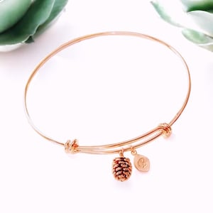 Könnte beinhalten: Ein roségoldenes, verstellbares Armkettchen mit einem kleinen Tannenzapfen-Anhänger und einem Buchstaben-Anhänger.