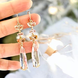 May include: A pair of gold-tone drop earrings featuring a vertical arrangement of faceted crystals in varying shapes, including small rectangular and round stones, culminating in a large, clear, teardrop-shaped crystal at the base of each earring.
