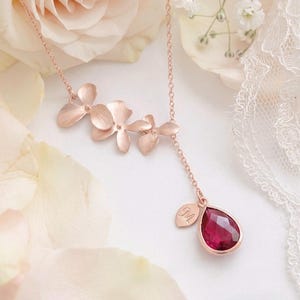 May include: A rose gold necklace featuring three flower pendants and a teardrop-shaped red gemstone. A small leaf charm with the letter "M" hangs near the gemstone. The necklace is displayed on a white surface with rose petals.