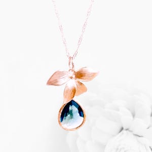 May include: A gold-toned pendant necklace featuring a floral design with a teardrop-shaped, blue gemstone. The pendant has a flower with three petals above the gemstone. The necklace is on a white background.