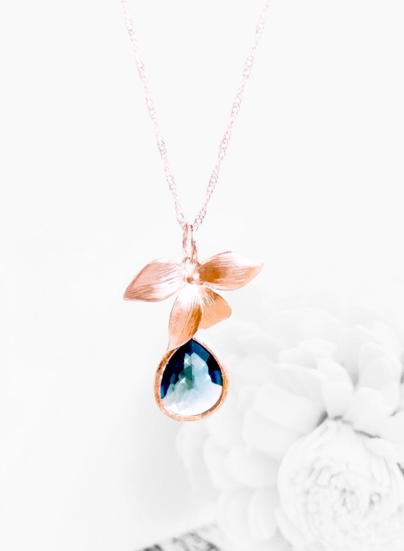 May include: A gold-toned pendant necklace featuring a floral design with a teardrop-shaped, blue gemstone. The pendant has a flower with three petals above the gemstone. The necklace is on a white background.