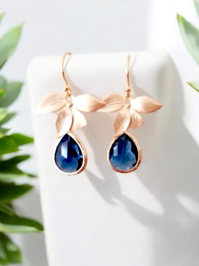 May include: A pair of rose gold earrings with a floral design. Each earring features three delicate petals and a teardrop-shaped, deep blue gemstone. The earrings are displayed on a white surface, with green foliage in the background.