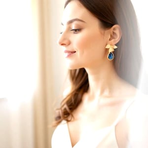 May include: Gold-toned leaf earrings with a teardrop-shaped, deep blue gemstone. The earrings are worn by a person with long, wavy brown hair and a white top. The earrings are the focal point, showcasing a delicate floral design.
