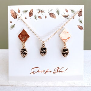 May include: A gold necklace and earrings set featuring a pine cone design. The necklace and earrings have a hammered gold finish and are displayed on a white card with a pine cone pattern. The card also has the text "Just for You!" printed on it.