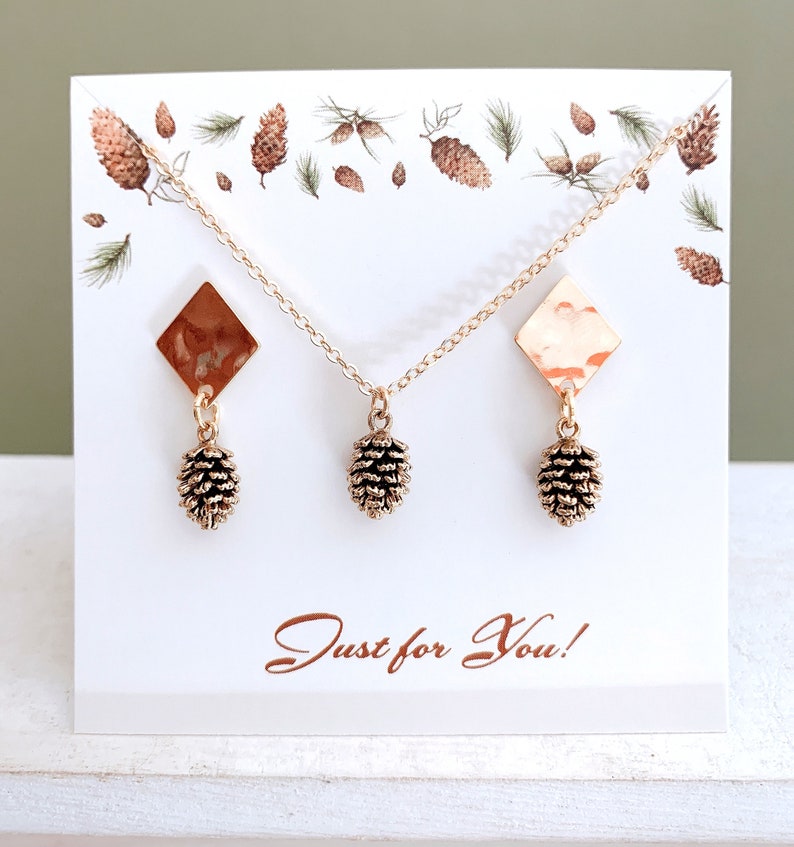 May include: A gold necklace and earrings set featuring a pine cone design. The necklace and earrings have a hammered gold finish and are displayed on a white card with a pine cone pattern. The card also has the text "Just for You!" printed on it.