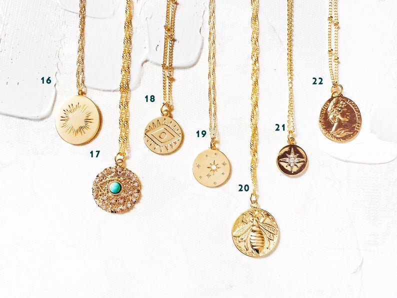 May include: Ten gold necklaces with different pendants, including a sun, an eye, a crescent moon, a star, a compass, a bee, and a profile of a woman. The necklaces are displayed on a white background.