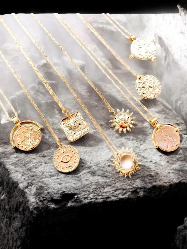 May include: A set of eight gold-toned necklaces with various pendant designs, including a sun, an eye, and a square. The necklaces are displayed on a gray textured surface.
