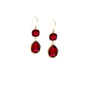 May include: A pair of gold-colored drop earrings with two red, gem-like stones. The top stone is round with a scalloped edge, and the bottom stone is a teardrop shape. The earrings hang from a gold-colored hook.