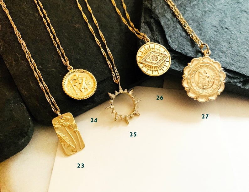 May include: Five gold necklaces with different pendants. The pendants include a sun, a flower, a face, an eye, and a religious symbol. The necklaces are on a dark background.