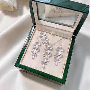 May include: A silver-colored jewelry set, including a necklace and earrings, displayed in a green box with a clear lid. The jewelry features a floral design. The box is lined with a cream-colored fabric.