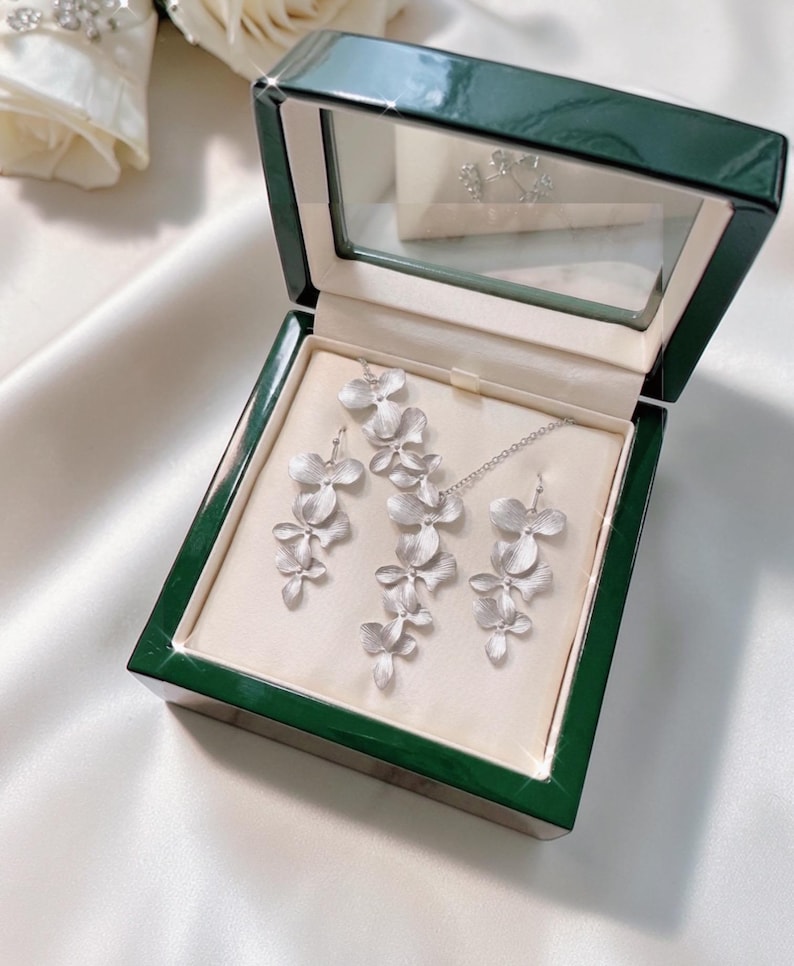 May include: A silver-colored jewelry set, including a necklace and earrings, displayed in a green box with a clear lid. The jewelry features a floral design. The box is lined with a cream-colored fabric.