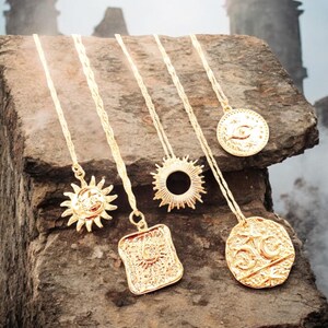 May include: Five gold necklaces with different pendants, including a sun, an eye, and a crescent moon, displayed on a rough stone surface.