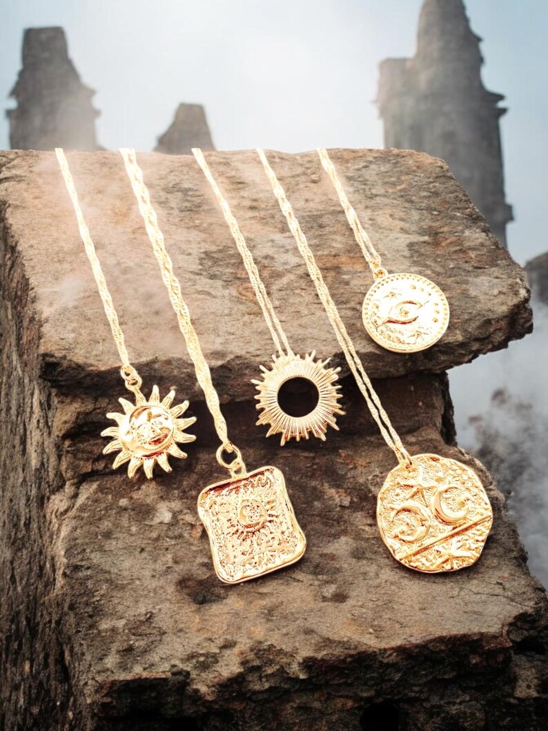 May include: Five gold necklaces with different pendants, including a sun, an eye, and a crescent moon, displayed on a rough stone surface.