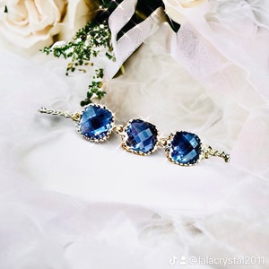 May include: A delicate gold chain bracelet with three square-shaped blue crystal stones. The bracelet is on a white fabric background.