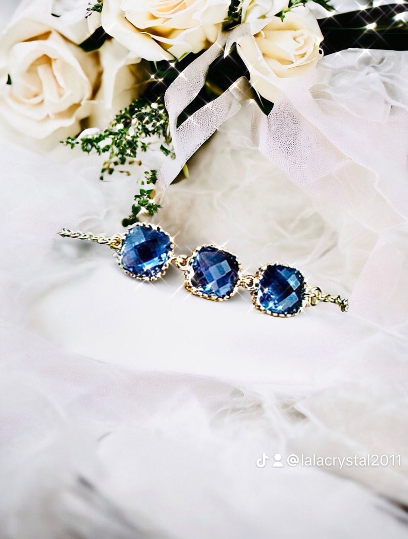 May include: A delicate gold chain bracelet with three square-shaped blue crystal stones. The bracelet is on a white fabric background.