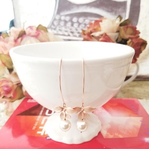 May include: A pair of delicate, rose gold earrings with a floral design and dangling faux pearls. The earrings are displayed on a white, seashell-shaped holder, set against a backdrop of a white bowl and a red book.